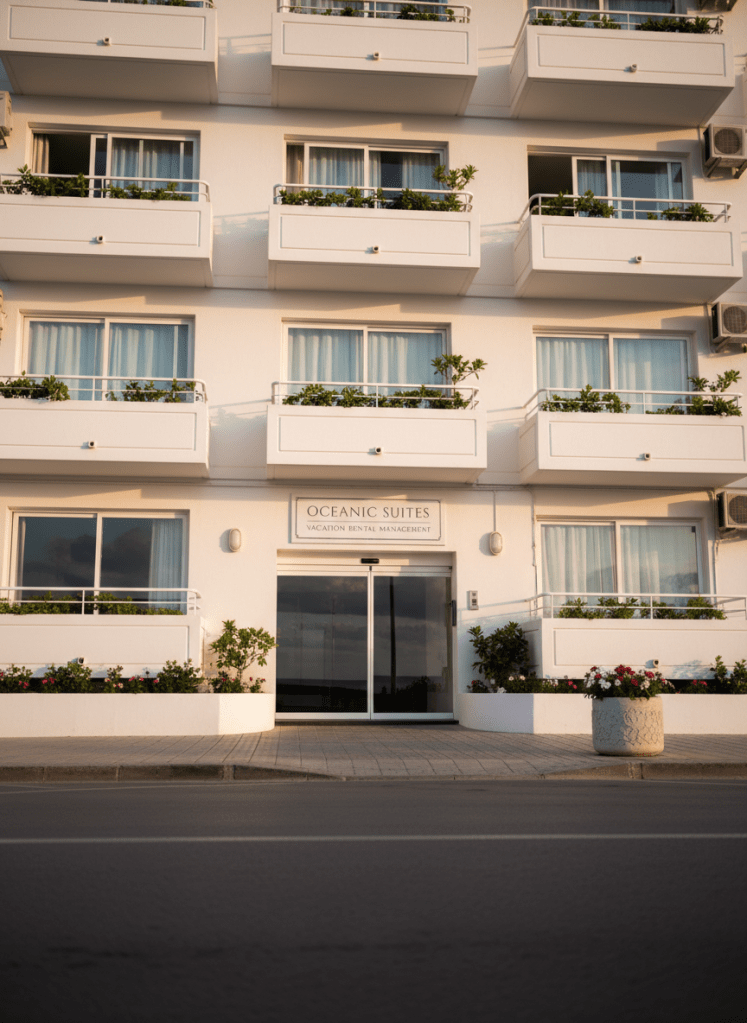 Una fachada impecable de un edificio de apartamentos turísticos en una zona costera, con balcones blancos alineados y plantas verdes bien cuidadas, mientras en la entrada se ve claramente un pequeño rótulo elegante con el nombre de una empresa de gestión vacacional. El entorno muestra aceras limpias, maceteros decorativos y una puerta de cristal brillante que refleja el cielo azul. La escena está iluminada por luz de mañana con un ligero brillo dorado, generando contrastes suaves y un ambiente optimista y confiable. Fotografía en perspectiva ligeramente angular desde la calle, con enfoque nítido en la entrada y profundidad hacia el resto del edificio, estilo fotográfico realista y profesional que transmite solidez y buena administración de propiedades.